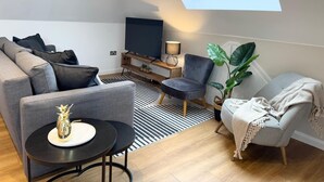 En moderne stue med en grå sofa, et rundt bord, et fjernsyn på et træstel og en stor plante.