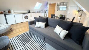 En moderne loftlejlighed med en grå sofa, et stribet tæppe og et køkken med hvide skabe og en vaskemaskine.