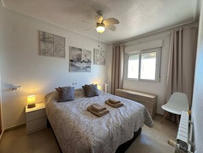 Une chambre à coucher comprenant un lit, des tables de chevet, un ventilateur de plafond, des œuvres d’art accrochées au mur, une fenêtre avec des rideaux et un radiateur.