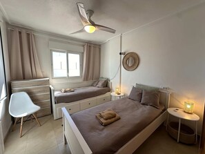 Ein Schlafzimmer mit einem Bett, zwei Nachttischen, einem Schreibtisch, einem Sessel, einem Deckenventilator und einem Fenster mit Vorhängen.