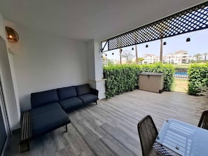 Ein überdachter Außenbereich mit einer Couch und Sesseln, einer Holzterrasse und Blick auf Gebäude im Hintergrund.