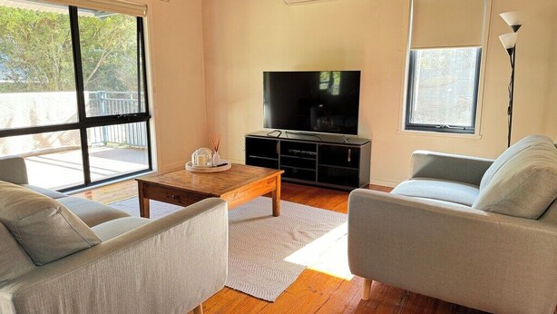 A living room with a flat-screen TV, two grey sofas, a wooden coffee table, and a large window with a view of trees. - Newly renovated, 3 bedroom, pet friendly unit. (Lorne)