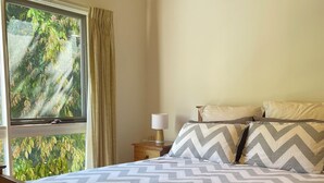 A bedroom with a wooden bed frame, a patterned bedspread, a bedside table with a lamp, and a window with curtains. - Newly renovated, 3 bedroom, pet friendly unit. (Lorne)