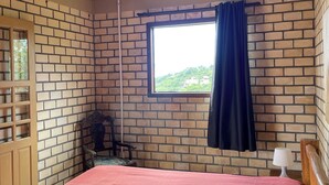 A room with a brick wall, a wooden ceiling, a bed with a red bedspread, a window with a view of greenery, and a hanging light fixture. - Hamarhavida (Imbituba)