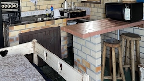 A kitchen with a brick backsplash, a gas stove, a microwave, and a sink. There are stools and a table with a lace tablecloth. - Hamarhavida (Imbituba)