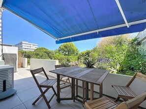 Un espace patio avec une table et des chaises, un auvent bleu et une vue sur la verdure et les bâtiments.