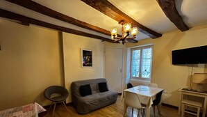 En hyggelig stue med trægulv, en grå sofa, en lille rund stol, et bord med stole, en lysekrone, et billede, et vindue og et fjernsyn.