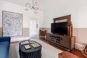 A living room with a large abstract painting, a blue sofa, a black coffee table with books, a wooden TV stand, and a wicker chair. - Casa Azula (Atlanta)