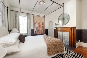 Une chambre à coucher avec un lit à baldaquin, une grande peinture abstraite, un miroir rond et une vue sur l’extérieur par une fenêtre.