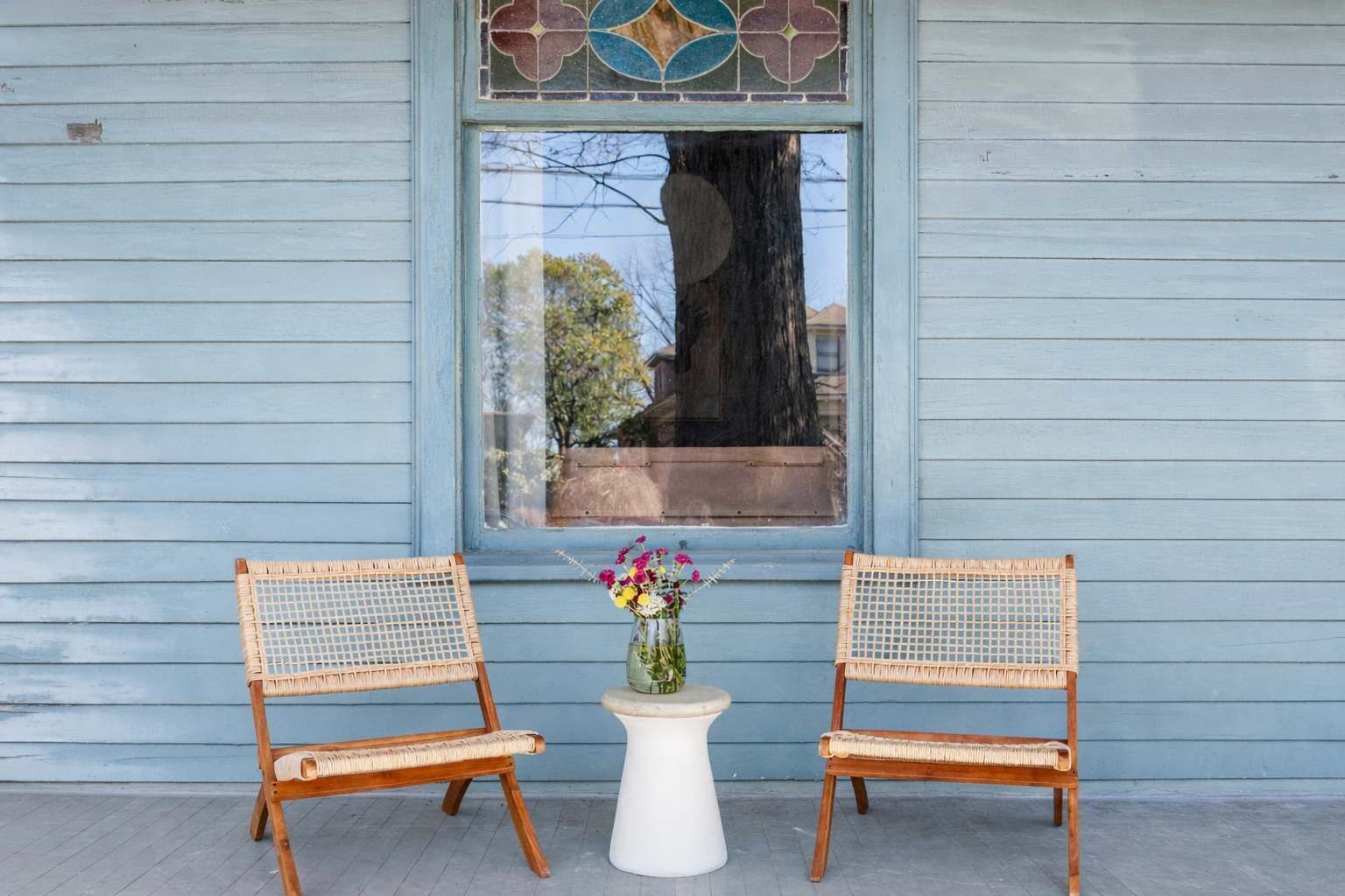 Dos sillas de mimbre con una mesita blanca entre ellas, ubicadas en un porche con un muro de madera azul y una ventana de vitral arriba.