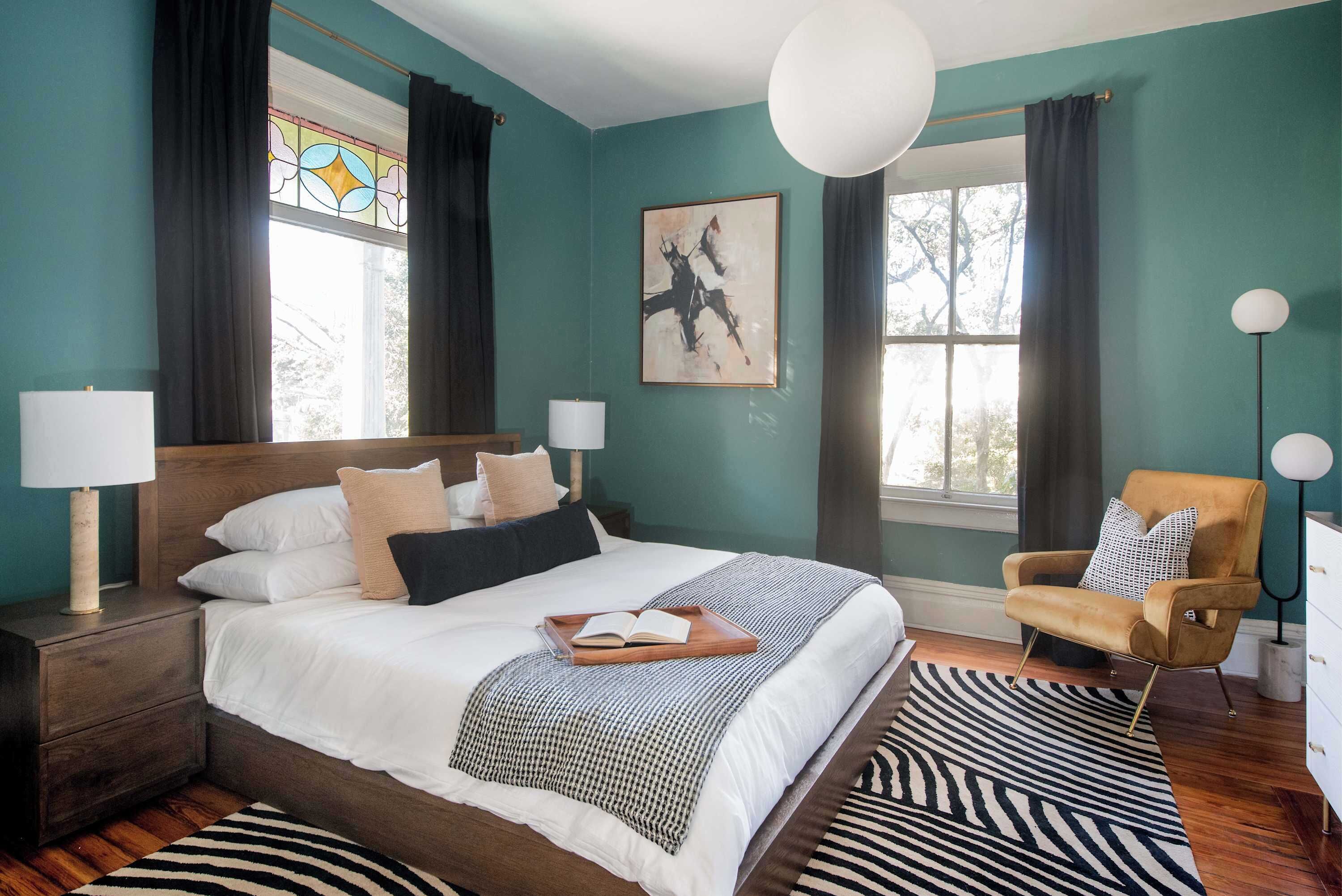A bedroom with a large bed, two bedside tables with lamps, a chair, a zebra-patterned rug, and a stained glass window.