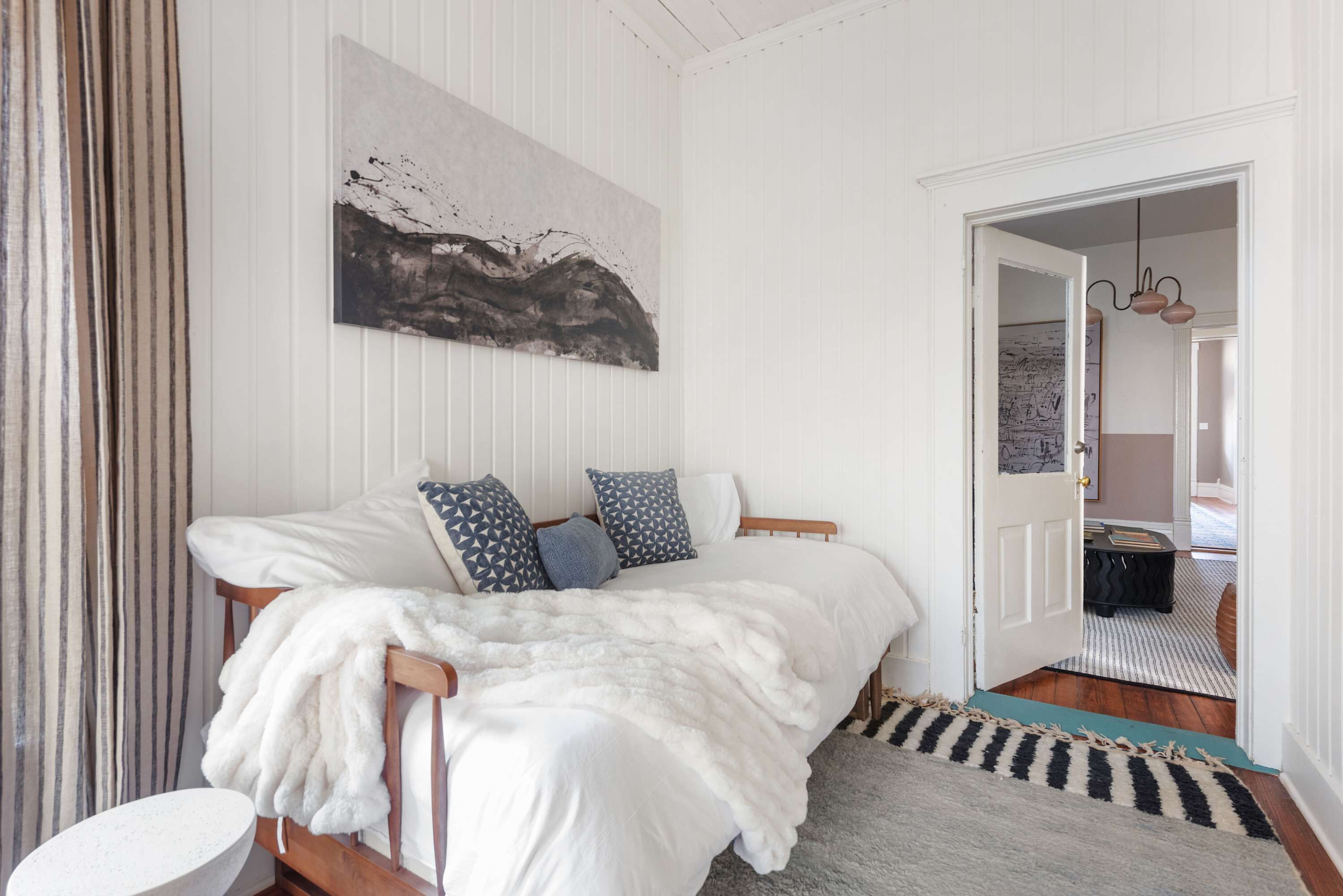 Ein Schlafzimmer mit einem Bett, einer weißen Wand, einem Gemälde und einer Tür zu einem anderen Raum.