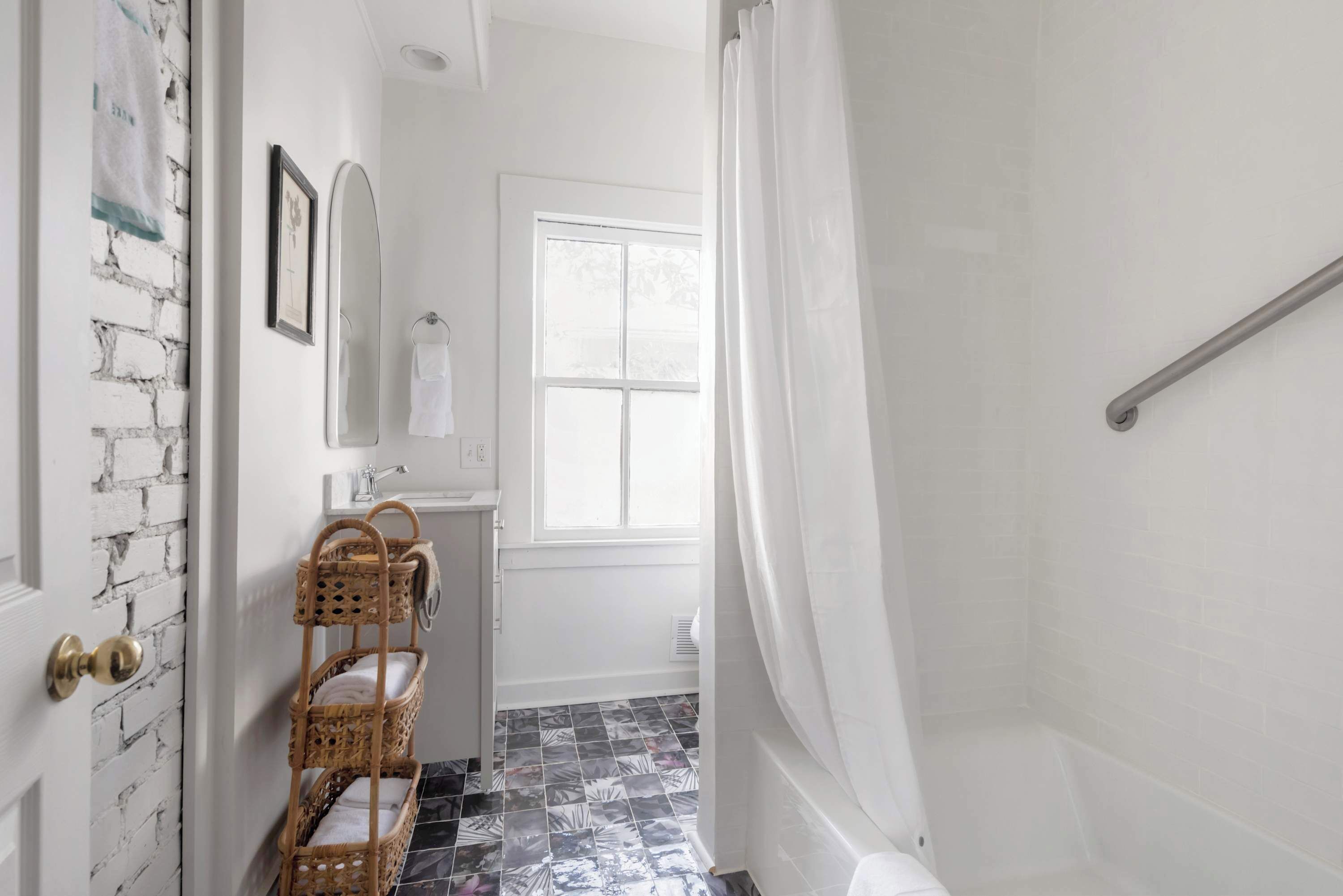 A bathroom with a white brick wall, a window, a shower with a curtain, a sink with a mirror, and a wicker shelf with towels.