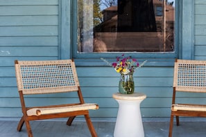 Two wicker chairs with a white table between them, placed on a porch with a blue wooden wall in the background.
