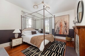 A four-poster bed with a beige throw blanket, a bedside table with a lamp, a black and white striped rug, and a large abstract painting on the wall. - Casa Azula (Atlanta)