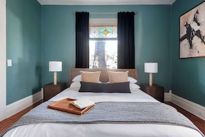 Ein Schlafzimmer mit türkisfarbenen Wänden, einem großen Bett mit weißer Bettwäsche, zwei Nachttischen mit Lampen, einem Buntglasfenster und einem Gemälde an der Wand.
