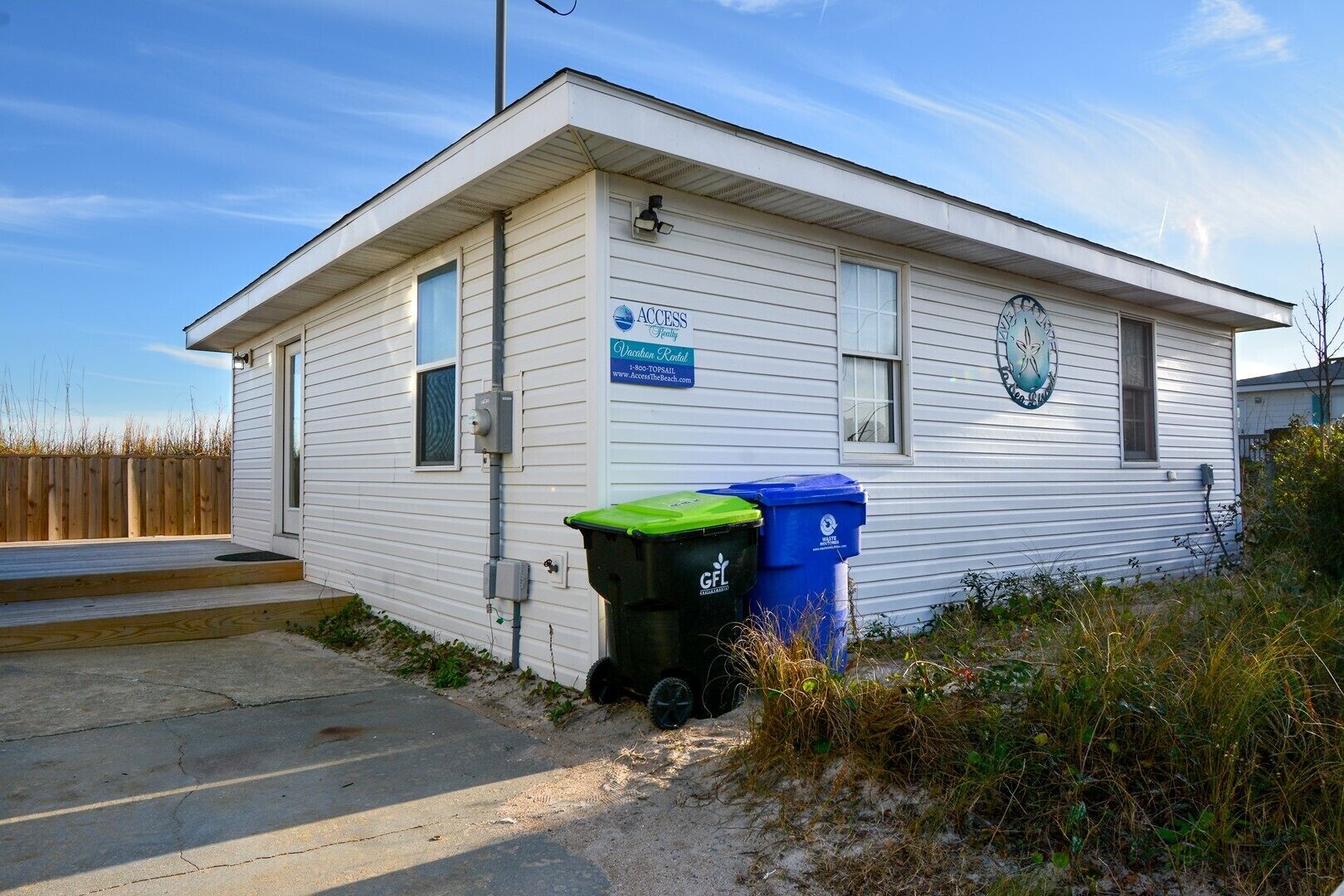 A single-story building with a sign indicating it is a vacation rental. There are two trash bins outside, one green and one blue, with recycling symbols.