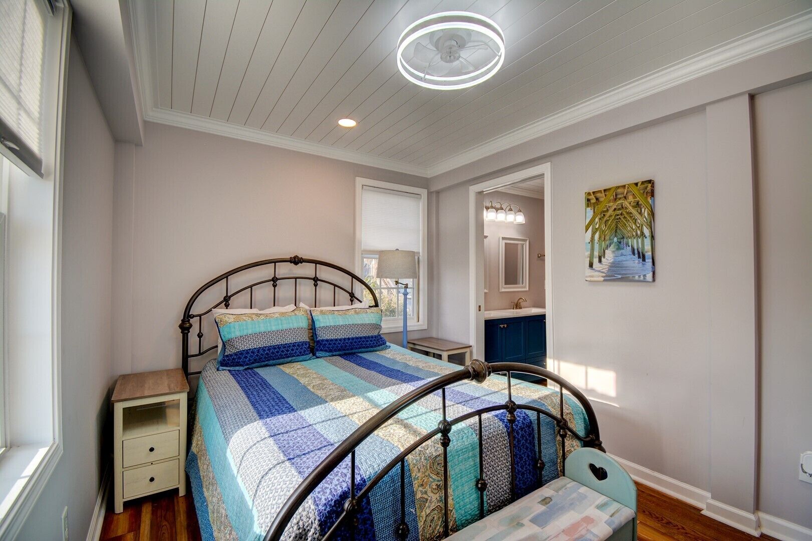 A bedroom with a bed, bedside table, and a nightstand. There is a window with blinds, a ceiling light, and a painting on the wall.