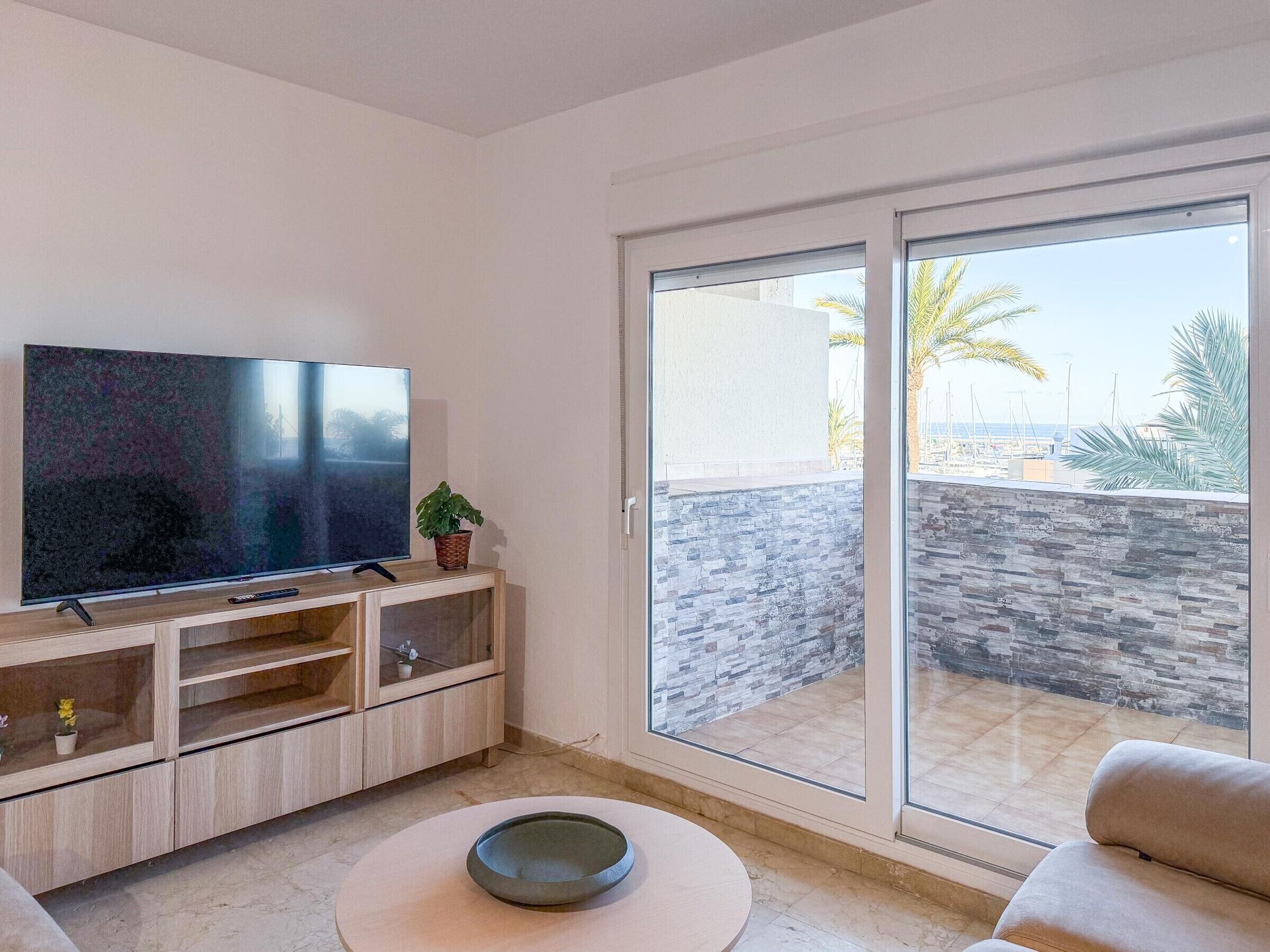 Un salon moderne avec un téléviseur à écran plat posé sur un meuble en bois, une table basse en verre et une porte-fenêtre coulissante donnant sur un balcon doté d’un mur en pierre et offrant une vue sur la mer.