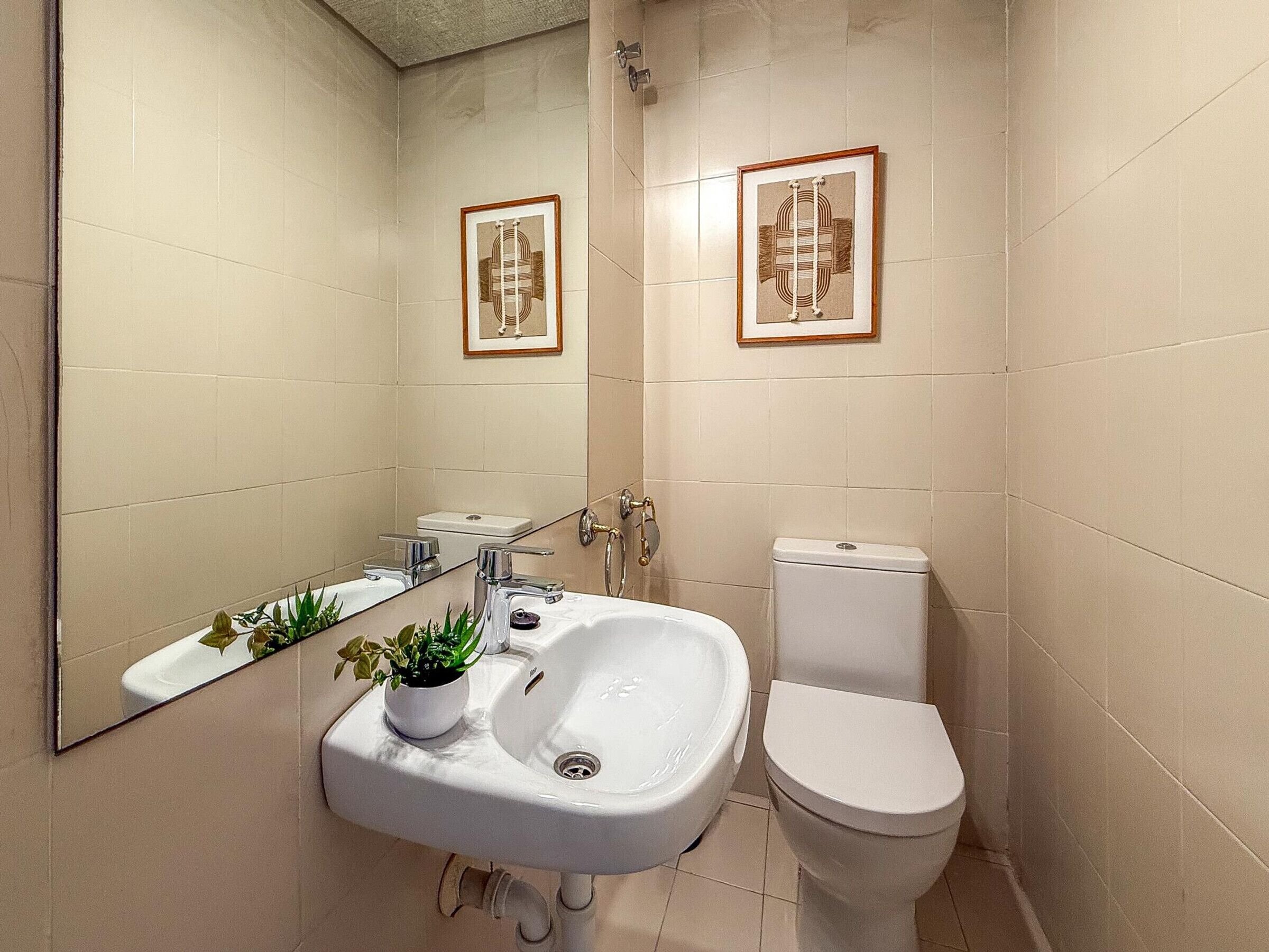 Un baño con un lavamanos blanco, inodoro y un espejo grande. Hay cuadros enmarcados en la pared y una planta pequeña sobre el lavamanos.