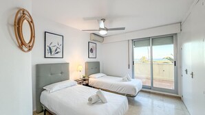 Una camera d'albergo con due letti, una ventola a soffitto, un'ampia finestra vista spiaggia e opere d'arte alle pareti.