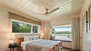 Un dormitorio con una cama grande, dos mesitas de noche, un ventilador de techo y un ventanal con vistas a un lago.