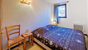 Ein Schlafzimmer mit Bett, Sessel, Nachttisch, Wandlampe und einem Fenster mit Vorhängen.