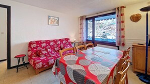 Ein Essbereich mit einer rot gemusterten Couch, einem Tisch mit geometrischem Tischläufer, Holzstühlen, einer Uhr und einem Fenster mit Blick auf Gebäude.