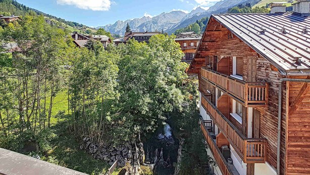 A wooden chalet with balconies, surrounded by lush greenery and mountains in the background.