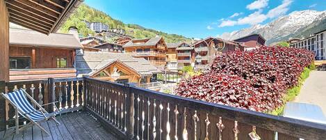 A wooden deck with a striped chair, a view of a mountain village with traditional buildings, and a red-leaved plant.