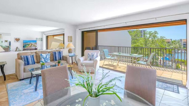 A modern living room with a glass table, two chairs, a sofa with blue and white cushions, and a balcony with a railing.