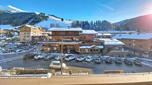 Eine schneebedeckte Stadt mit einem Parkplatz voller Autos, Gebäuden mit Balkonen und einem Berg im Hintergrund.