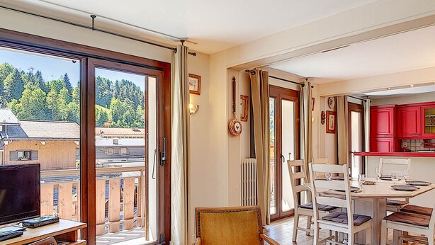 Ein Zimmer mit einem Holztisch, einem Stuhl, einem Esstisch mit Stühlen und Blick auf Gebäude und Bäume durch große Fenster.