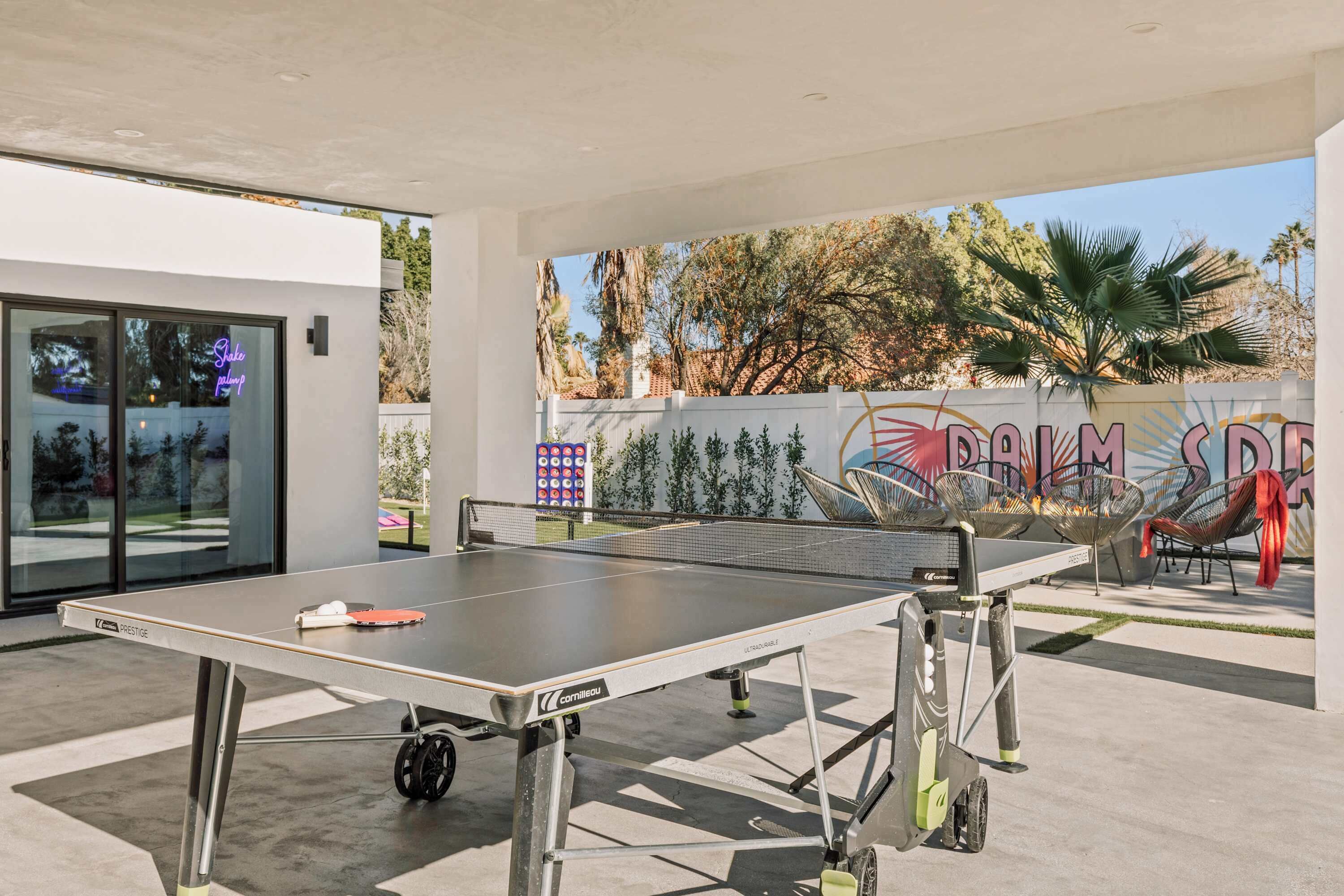 An outdoor ping pong table with a neon sign, a mural, and a palm tree.