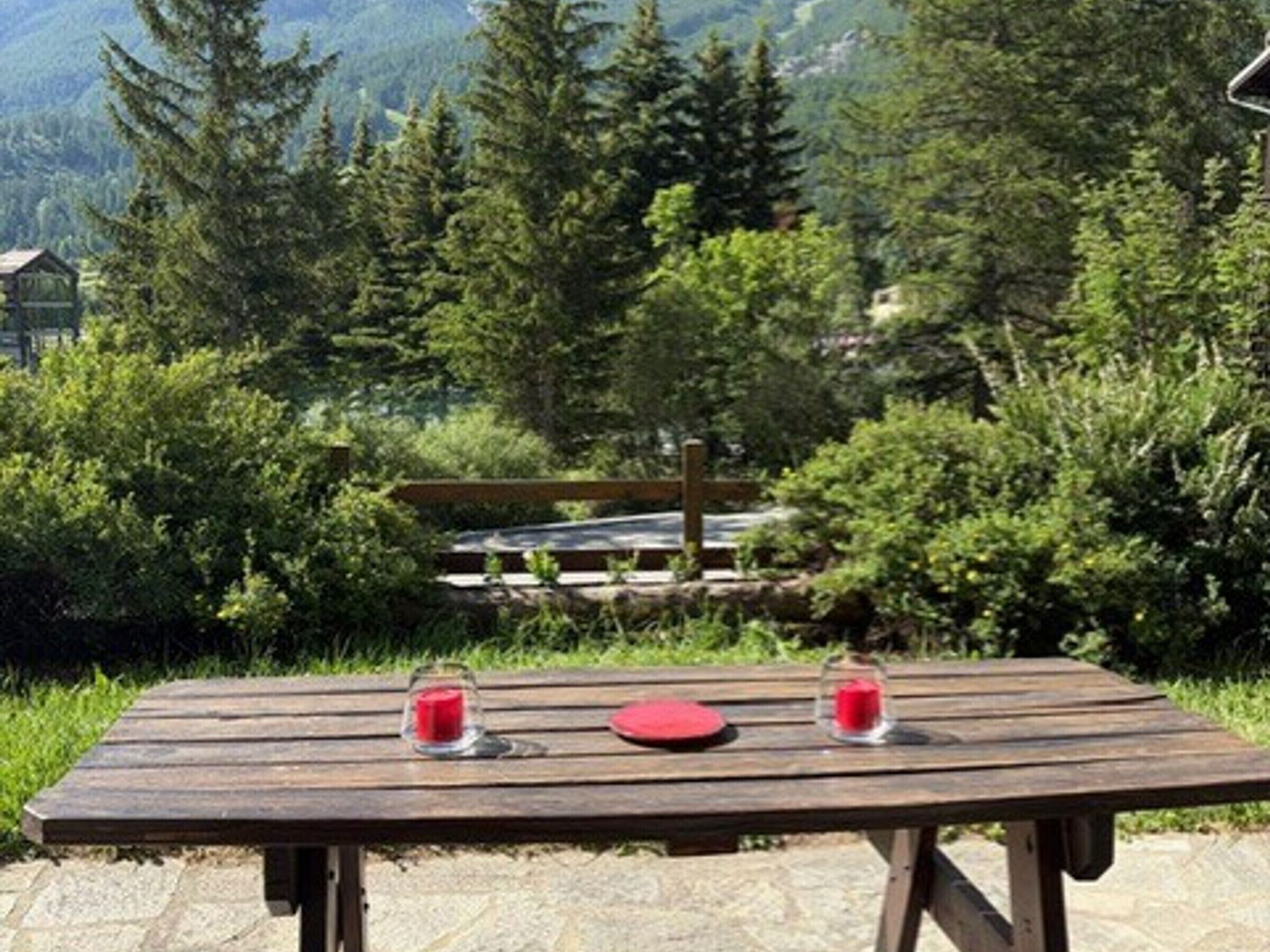 A wooden picnic table with two glasses of red drink and a red plate, set against a backdrop of lush greenery and distant mountains.