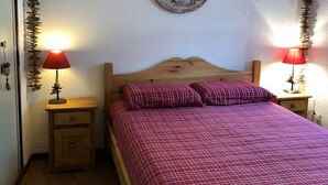 Un dormitorio con cama de madera, ropa de cama a cuadros rojos, dos mesitas de noche con lámparas y un cuadro en la pared que representa una escena histórica.