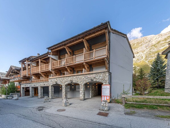 A traditional chalet with wooden balconies and stone pillars, set against a mountainous backdrop.