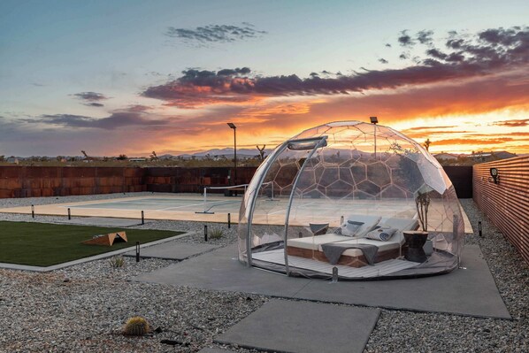A geodesic dome structure with a seating area inside, surrounded by a paved patio and a view of a sunset.