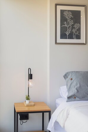 Private bedroom corner with crisp white bedding, a soft gray accent pillow, botanical artwork, and a warm bedside lamp. - Comfortable Studio Apartment in Nunoa Ideal for 2 (Ñuñoa)
