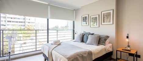 Bright studio sleeping area with a neatly dressed bed, botanical artwork, and expansive windows overlooking the city and surrounding trees.