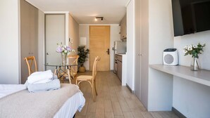 A hotel room with a bed, a desk, a chair, a small table with a vase of flowers, a television mounted on the wall, and a kitchenette area. - Chic Urban Studio Perfect for Business or Leisure (Ñuñoa)
