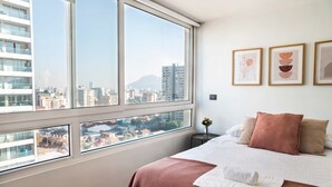 Une chambre avec une grande fenêtre, un lit recouvert d’une couverture blanche et marron, et trois œuvres d’art encadrées accrochées au mur.