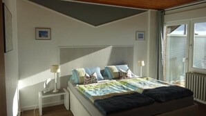 A bedroom with a bed, bedside tables, a nightstand, a window with blinds, and a ceiling with wooden panels. - Hotel Miramar  (Dahme)