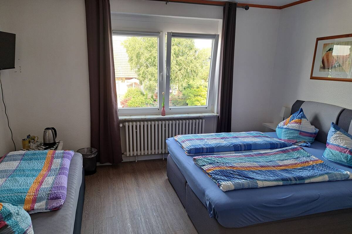 Une chambre avec deux lits, un radiateur, une fenêtre avec des rideaux, une petite table avec une bouilloire et un tableau encadré au mur.