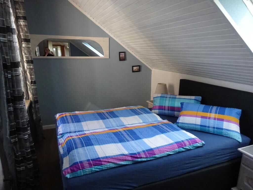 Un dormitorio con techo inclinado, una cama con colcha escocesa, dos almohadas, una mesita de noche y un espejo.