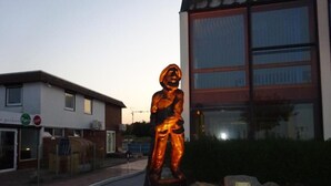 A statue of a person in a suit, placed on a stone pedestal with rocks around it, in front of a building with large windows. - Hotel Miramar  (Dahme)