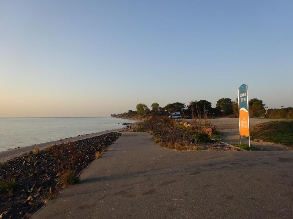 Uma passarela pavimentada ao lado da água, um sinal indicando área de estacionamento, e uma área gramada com árvores ao fundo.