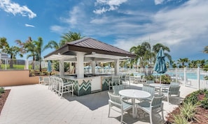 A covered outdoor seating area with tables and chairs, a pool, and palm trees in the background.