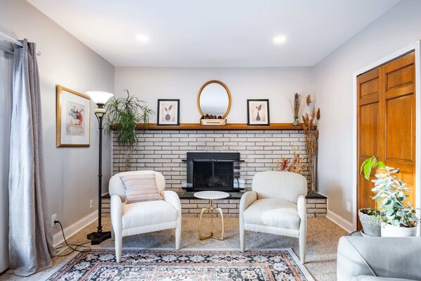 A living room with a fireplace, two white armchairs, a small round table, and a patterned rug.