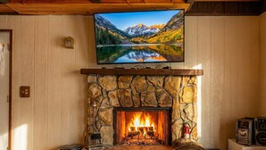 A fireplace with a stone surround, a television mounted above it, and a fire burning inside. -   ‼️Comfy 2BR Getaway Near Lake Grego w/ Mtn Views (Crestline)
